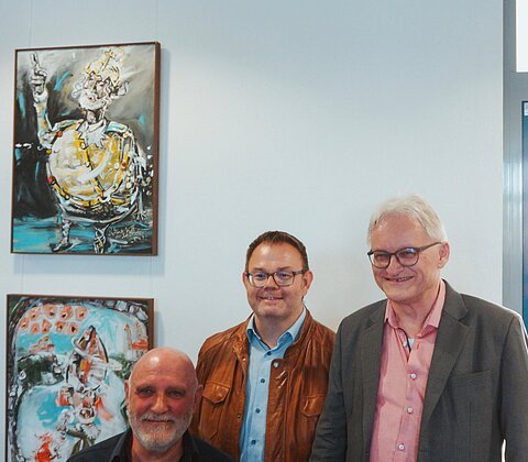 Künstler Uli Koch, Bürgermeister Dr. Benedikt Grünewald und Archivar Dr. Georg Köglmeier stehen gemeinsam bei der feierlichen Eröffnung der Ausstellung. In entspannter Atmosphäre tauschen sie sich vor einem Kunstwerk aus – ein Moment, der Kunst, Geschichte und Gemeindeleben verbindet.