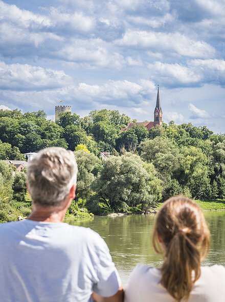 Ein Mann und eine Frau sind von hinten zu sehen, wie sie auf die Donau blicken. Das Donauufer ist mit bäumen begrünt und im Hintergrund spitzen der Heinrichsturm und die Kirche St. Nikolaus aus grünen Baumkronen unter bewölktem Himmel.