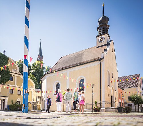 Vier erwachsene stehen in der Marktmitte neben der Marktkirche und folgen einem Gästeführer. Der Markt ist mit rot gelben Wimpeln dekoriert und links steht der blau-weiß gestreifte Maibaum steht unter blauem Himmel.