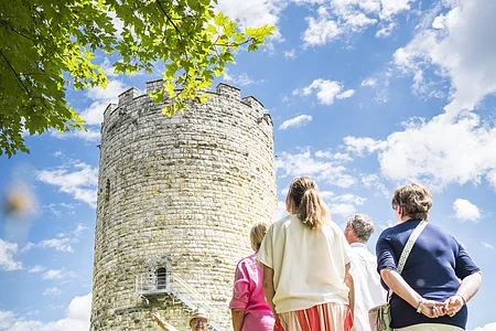 Vier Erwachsene nehmen an einer Marktführung vor dem Heinrichsturm teil. Der Himmel ist blau-weiß und oben spitzen grüne Äste in das Bild.