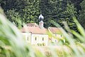Im Vordergrund ist unscharfes langes wehendes grünes Gras. Im Hintergrund ist die Kirche Frauenbründl zu sehen, welche am Rande eines Waldes steht. Sie ist gelb mit rotem Dach und einem kleinen Turm.