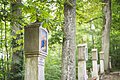 Zu sehen ist eine Station der Mariengrotte in Dünzling. Es handelt sich um eine Steinsäule mit einem Bild. Im Hintergrund sind drei weitere Stationen zu sehen. Diese stehen zwischen zwei Bäumen mit grünen Kronen.