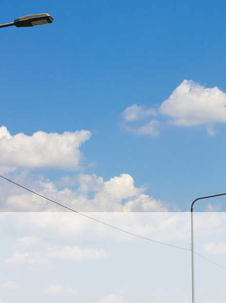 Zu sehen sind 3 Straßenlaternen vor blauem Himmel mit weißen Wolken. Die Laternen sind durch ein Kabel verbunden.
