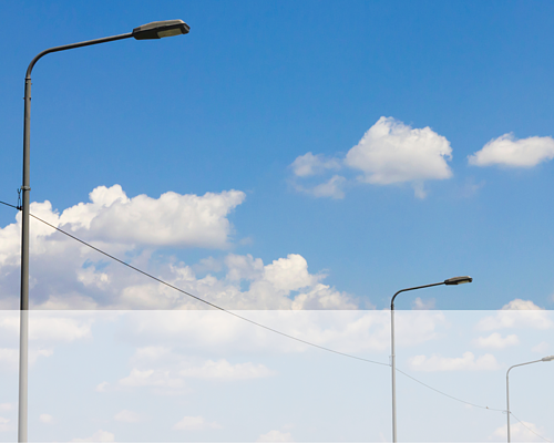Zu sehen sind 3 Straßenlaternen vor blauem Himmel mit weißen Wolken. Die Laternen sind durch ein Kabel verbunden.