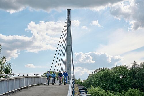 Eine vierköpfige Familie fährt über die Fußgängerbrücke in Bad Abbach in Richtung Kamera unter einem weiß-blauen Himmel.