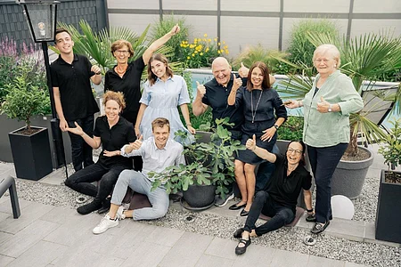 Gruppe von neun Personen steht und sitzt in einem Garten, alle zeigen Daumen hoch, umgeben von Pflanzen.