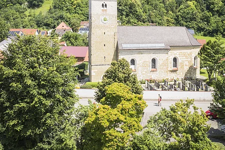 Das Luftbild zeigt die Kirche in Oberndorf aus erhöhter Perspektive. Die romanisch anmutende Kirche mit quadratischem Turm und rotem Zeltdach steht im Zentrum des Bildes. Umgeben ist sie von einem gepflegten Friedhof mit Grabsteinen und grünem Rasen. Der Himmel ist blau mit leichten Schleierwolken.