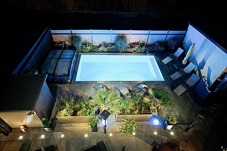 Beleuchteter rechteckiger Pool in einem Garten bei Nacht, umgeben von Pflanzen und Liegestühlen.