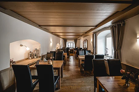Restaurant mit Holztischen und schwarzen Stühlen, Holzboden und Decke, Wandleuchten, Fenster mit Vorhängen.