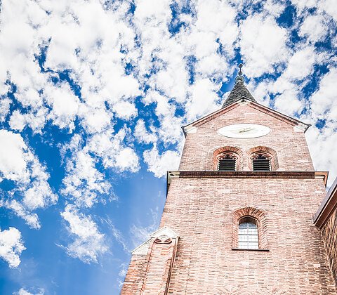 Zwei Personen mit Rucksäcken betrachten einen hohen, roten Backsteinkirchturm vor einem bewölkten Himmel.