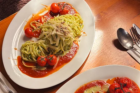 Zwei Teller mit grüner Pasta, Tomatensoße, Kirschtomaten und geriebenem Käse auf Holztisch.
