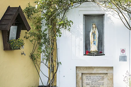 Steinstatue in einer Nische, umgeben von Rosenranken, mit einem kleinen Brunnen darunter und Blumen im Vordergrund.