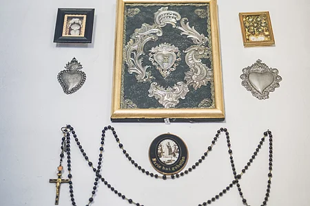 Wanddekoration mit mehreren gerahmten Bildern, zwei herzförmigen Objekten und einem Rosenkranz mit Kreuz.