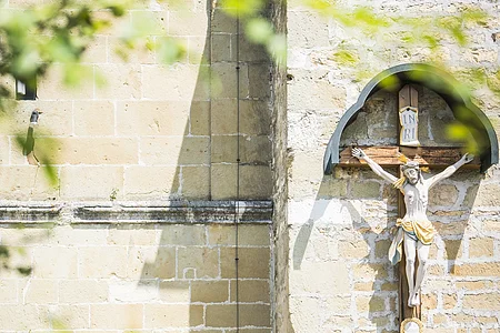 Zwei Wanderer gehen auf beiges Kirchengemäuer zu. An der Kirche hängt ein Jesuskreuz und eine braune alte Holztüre führt hinein. Es ist sonnig.