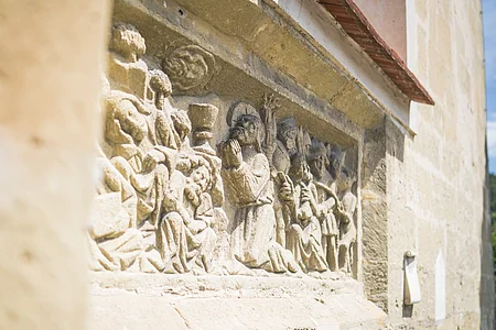 Steinrelief an einer Außenwand mit mehreren Figuren in historischer Kleidung, darunter eine betende Person.