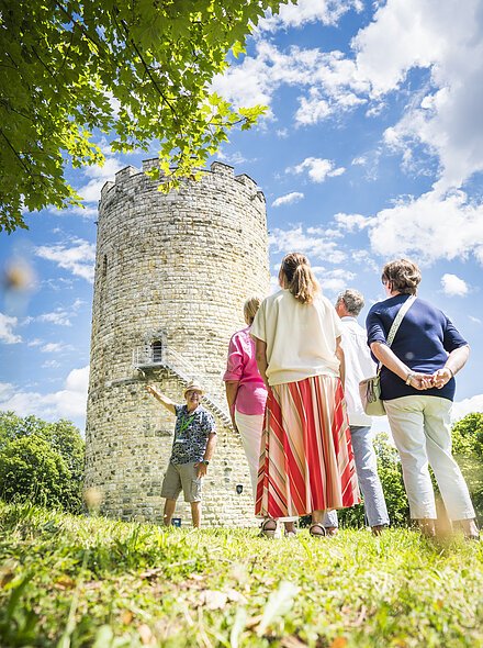Gruppe von Menschen steht vor einem alten runden Steinturm (Heinrichsturm) im Freien, ein Mann zeigt auf den Turm.