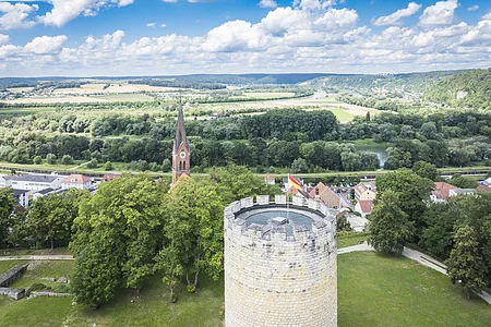 Steinturm mit Flagge, Kirche im Hintergrund, umgeben von Bäumen und Landschaft.
