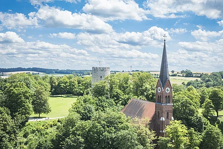 Nikolauskirche und Burgbergbbach Kirche mit rotem Dach und Turm mit Uhr in grüner Landschaft, daneben ein runder Steinturm.