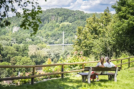 Blick vom Burgberg Ein Paar sitzt auf einer Bank mit Blick auf eine bewaldete Landschaft und eine Brücke im Hintergrund.