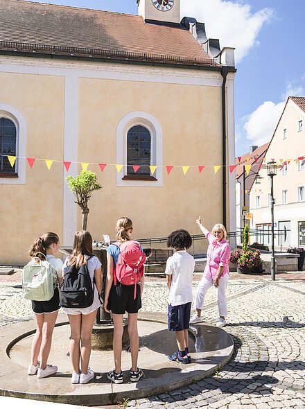 Kinderführung Marktmitte Eine Gruppe von Kindern steht um einen Brunnen, eine Frau zeigt auf eine Kirche mit Turm und Uhr.