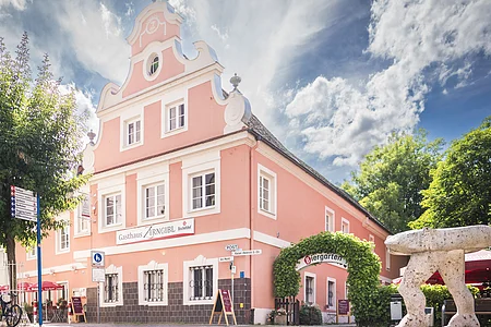 Ein rosa Gebäude mit Barockfassade, daneben ein Biergarten mit grüner Laube, im Vordergrund eine Steinfigur.