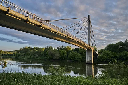 Fußgängerbrücke über einen Fluss, umgeben von Bäumen und bewölktem Himmel.