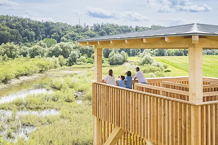 Eine vierköpfige Familie steht auf dem Vogelbeobachtungsturm - ein viereckiges Konstrukt aus Holz mit einem grauen Dach - und blicken in eine renaturierte Kiesgrube.