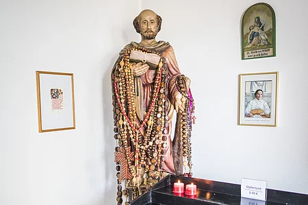 Statue mit Rosenkränzen behängt, daneben ein Kerzenhalter mit roten Kerzen und ein Schild mit "Opferlicht 0,50 €".