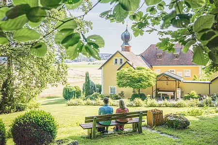 Zwei Personen sitzen auf einer Bank in einem Garten, im Hintergrund ein gelbes Gebäude mit Turm.