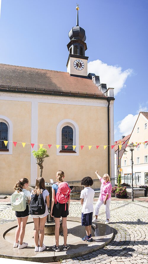 Eine Gruppe von Kindern steht um einen Brunnen, eine Frau zeigt auf eine Kirche mit Turm und Uhr.