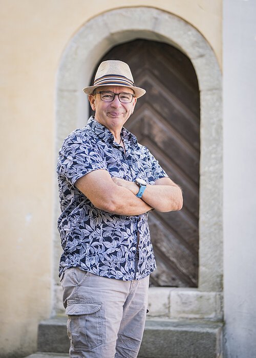 Gästeführer Portrait Eine Person mit Hut und Brille steht mit verschränkten Armen vor einer Holztür in einem steinernen Rahmen.