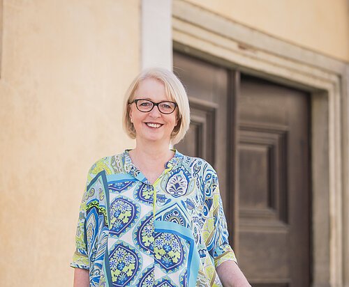 Gästeführer Portrait Eine Person mit Brille steht lächelnd an einem Geländer vor einer Holztür.