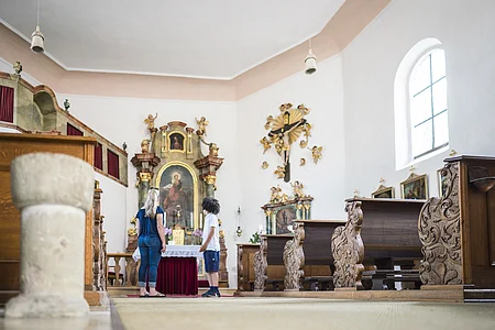 Zwei Personen stehen in einer Kirche vor einem Altar mit religiösen Kunstwerken und geschnitzten Holzbänken.
