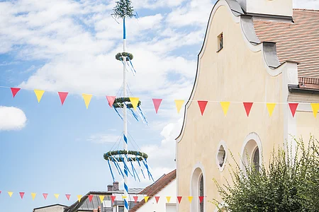 Stadtplatz mit Kirche, Maibaum und bunten Wimpeln; blauer Himmel im Hintergrund.