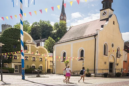 Ein Maibaum mit Kränzen steht auf einem Platz vor einer gelben Kirche, umgeben von bunten Wimpeln.