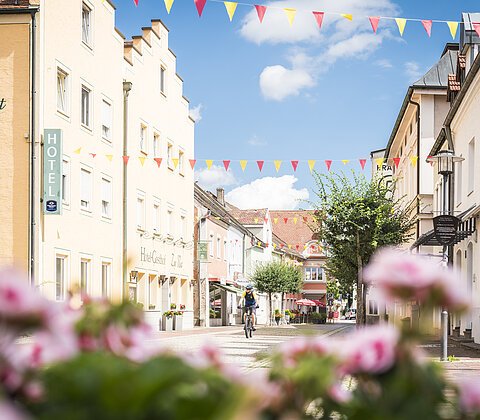 Ein Radfahrer fährt in der Marktmitte an dem Hotel Zur Post vorbei in Richtung Kamera. Der Himmel ist blau-weiß und im Vordergrund sind unscharf weiß-rosa Blumen.