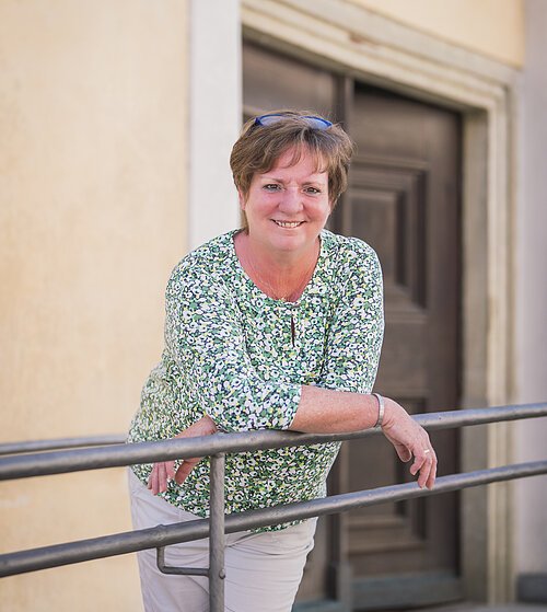 Gästeführer Portrait Eine Person lehnt sich an ein Geländer vor einer Tür, trägt ein grünes, geblümtes Oberteil.