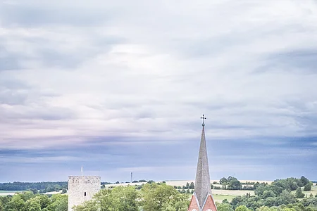 Luftaufnahme einer Kirche mit spitzem Turm, umgeben von Bäumen, im Hintergrund ein Turm und Felder.