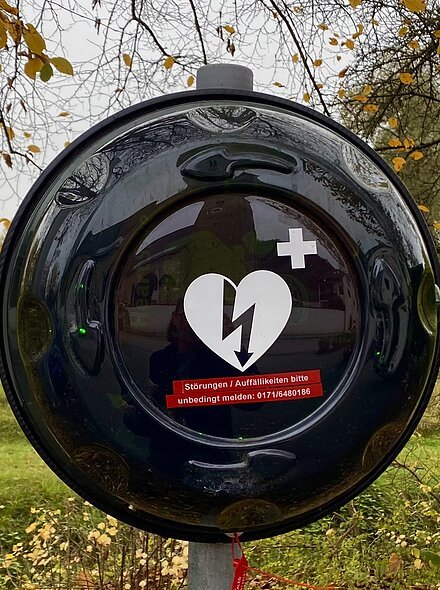 AED - Parkplatz Kirche Oberndorf Außen an einem Baum befestigtes schwarzes Gehäuse für einen Defibrillator im Herbst mit gelben Blättern.