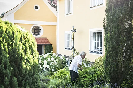 Das Bild zeigt einen gepflegten kleinen Garten mit blühenden Stauden und Buchsbäumen. Im Vordergrund steht ein älterer Mann, der sich um die Pflanzen kümmert. Im Hintergrund ist ein ockergelbes Gebäude mit weißen Fensterrahmen und einem kleinen Zwiebelturm zu sehen. An der Hauswand befindet sich ein schmiedeeisernes Grabkreuz mit goldenen Akzenten.