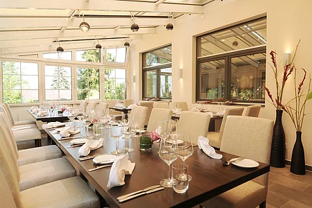 Hell eingerichteter Speiseraum mit gedeckten Tischen und großen Fenstern zu einem Garten hin.