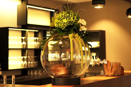 Große Glasvase mit grünen Blumen und Blättern auf einem Tresen in einem warm beleuchteten Raum.