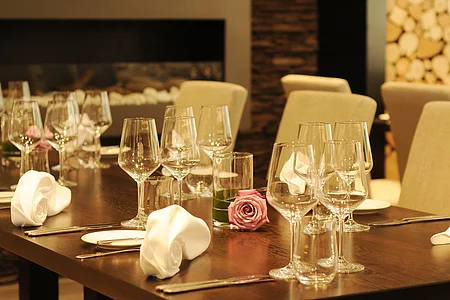 Esstisch mit eingedeckten Gläsern, weißen Servietten und einer rosa Rose in einem Glas auf dunklem Holz.