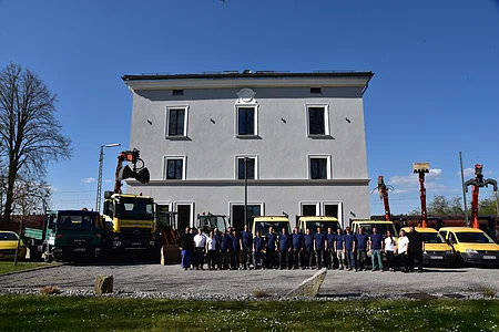 Gruppenfoto von 23 Personen vor einem weißen Gebäude mit mehreren gelben und grünen Baumaschinen und Fahrzeugen.