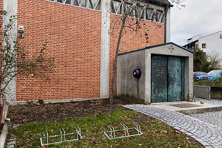 Fahrradständer auf Rasen vor roter Backsteinwand und kleinem Betongebäude mit Metalltür und Defibrillator.