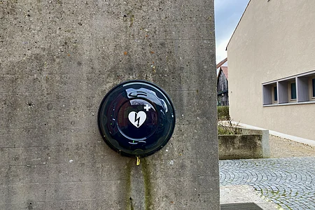 Defibrillator in schwarzem Gehäuse mit Herzsymbol an grauer Betonwand befestigt, daneben gepflasterter Weg und Gebäude.