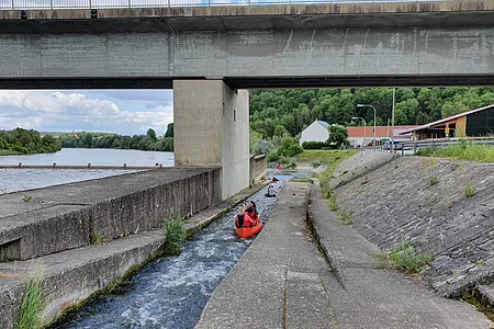 Menschen in roten Kajaks fahren auf einem schmalen Wasserkanal unter einer Brücke entlang.