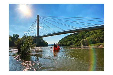 Personen paddeln in einem roten Kanu auf einem Fluss unter einer Hängebrücke bei sonnigem Himmel.