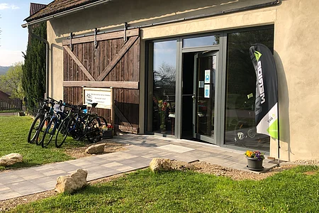 Eingang eines Gebäudes mit Fahrrädern, Holztür, Glasfenstern und Werbefahne auf einem gepflasterten Weg.