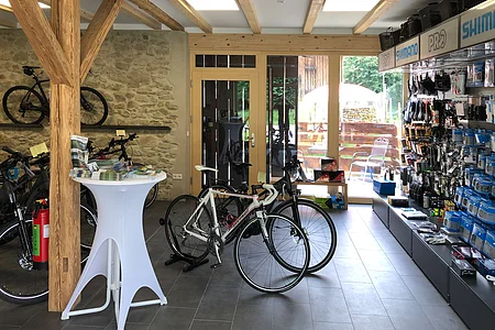 Innenraum eines Fahrradladens mit mehreren Fahrrädern, Holzstützen und Regalen mit Zubehör an der rechten Wand